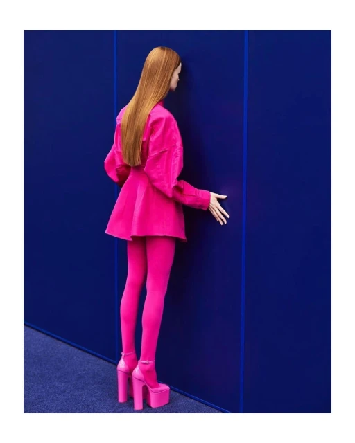 Woman in bright pink skirt suit and platform heels poses against a royal blue wall in a minimalist studio setting. Fashion photography.