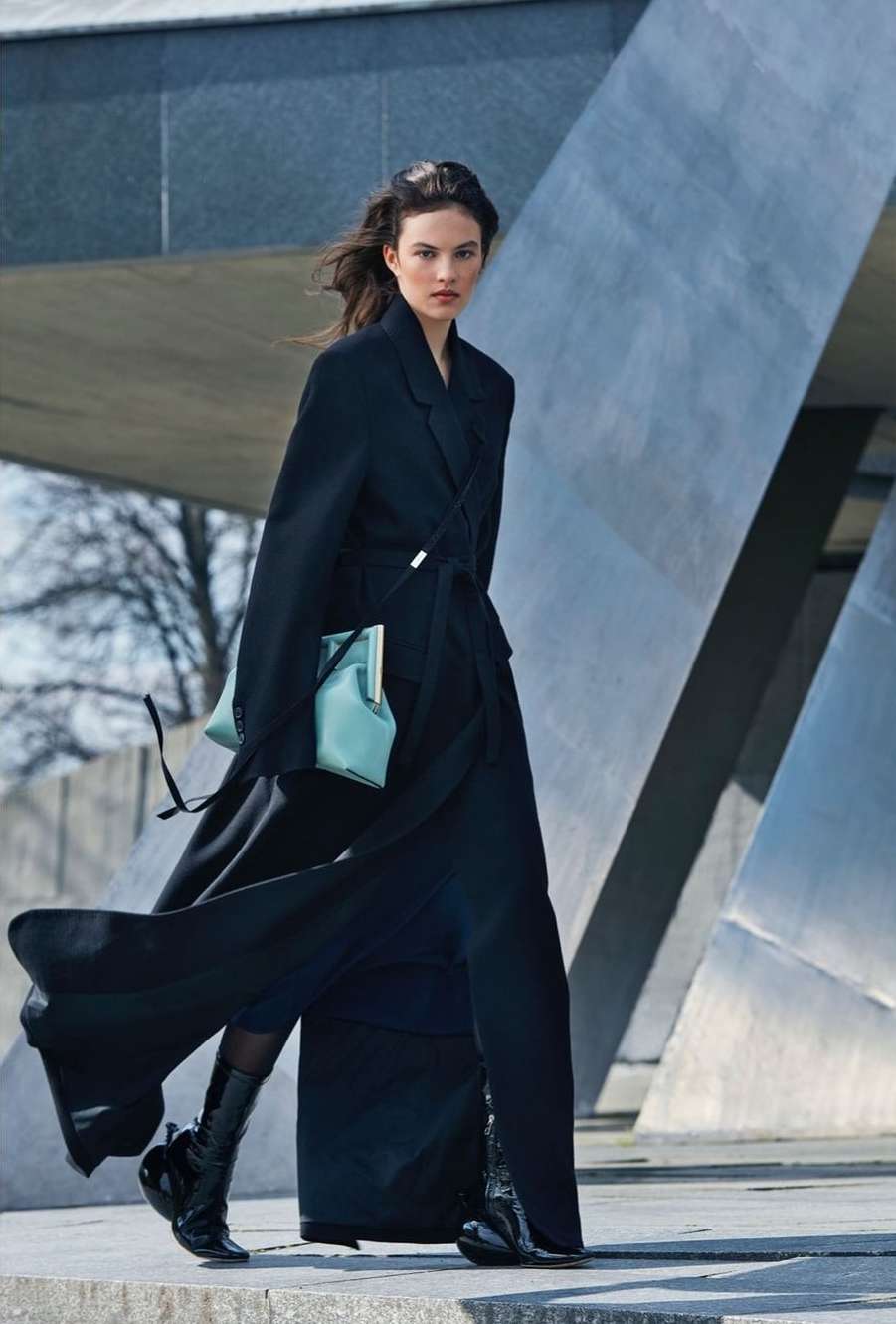 Model in a long black coat holding a light blue clutch walking outdoors near architectural concrete structures.