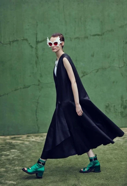 Fashionable woman with winged sunglasses and a long black dress walks on a green surface, creating a modern and stylish scene.