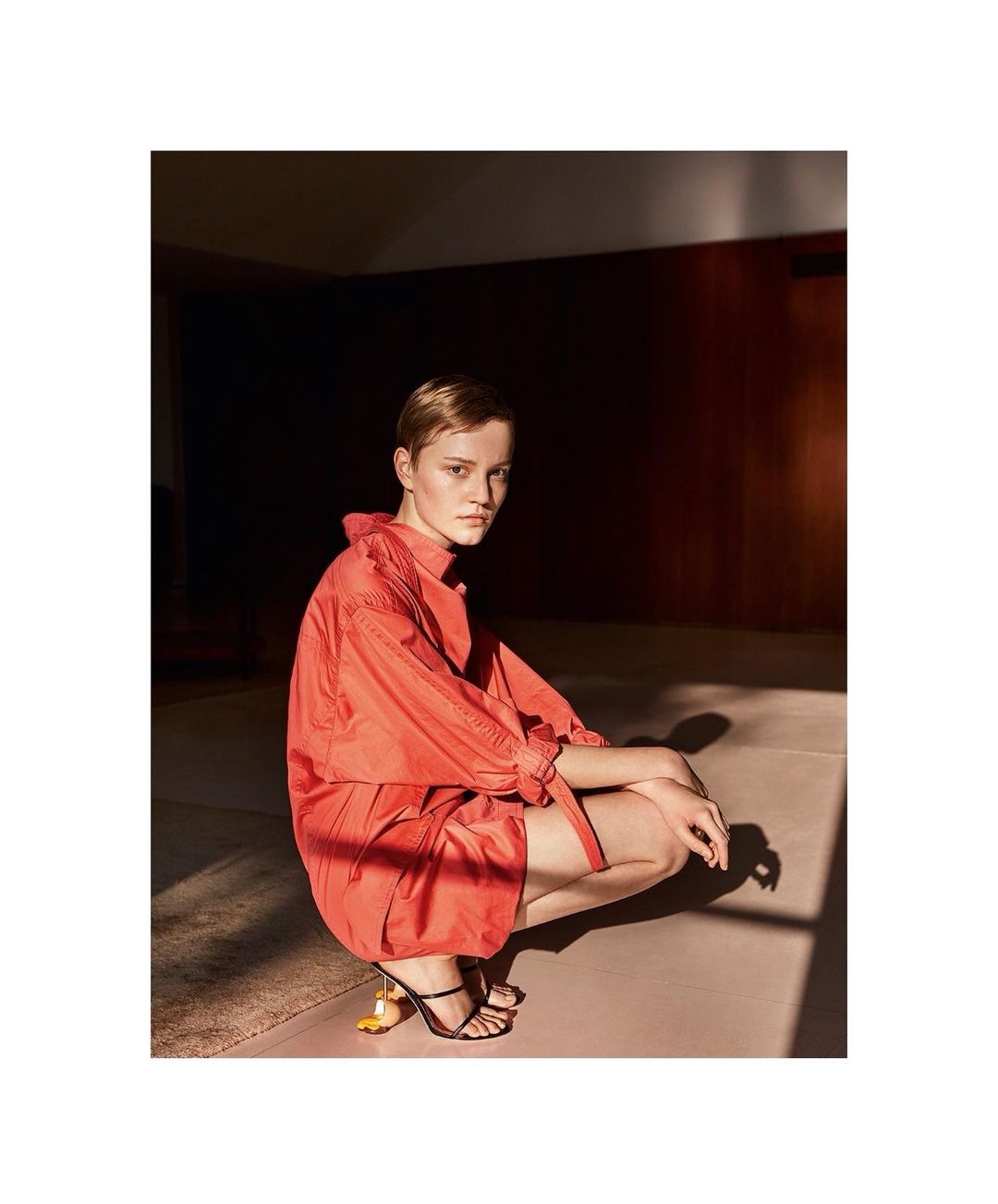 Fashion shot of a young adult woman in an orange shirt and shorts crouching indoors, highlighting modern style and a unique shoe design.