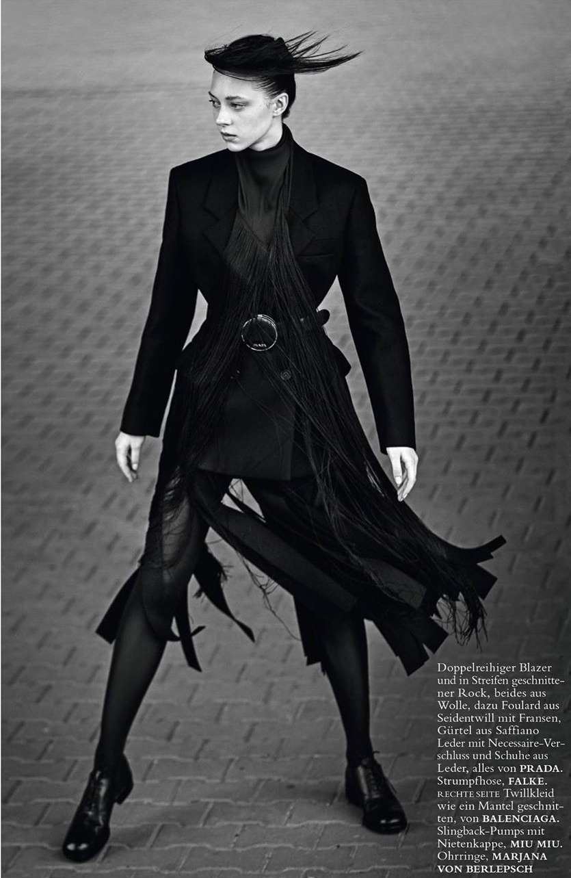 A woman in a black blazer, top, and pants with a fringed belt poses against a gray brick background in a fashion shot.