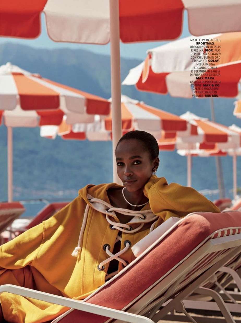 Woman in yellow hooded top lounges on a beach chair under an umbrella, with mountains visible in the distance on a sunny day.