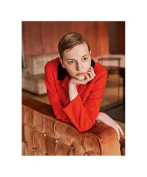 A woman with short hair wearing a red jacket leaning on a tufted velvet sofa indoors
