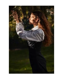 Profile view of a woman with long curly hair wearing a white shirt and black dress reaching for tree branches.