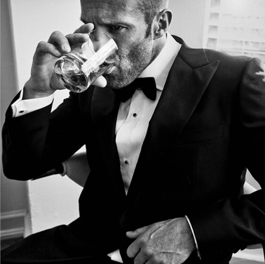 Jason Statham in tuxedo drinking from glass in a high-contrast black and white portrait, seated indoors.