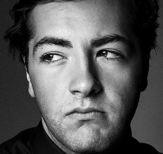 Black and white portrait of an adult male with medium length hair looking off to the side with a serious expression. 
