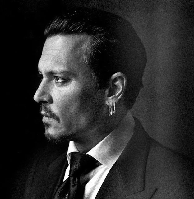 Black and white portrait of adult male wearing suit and tie, with dangling earring on dark gray backdrop, captured in studio lighting.