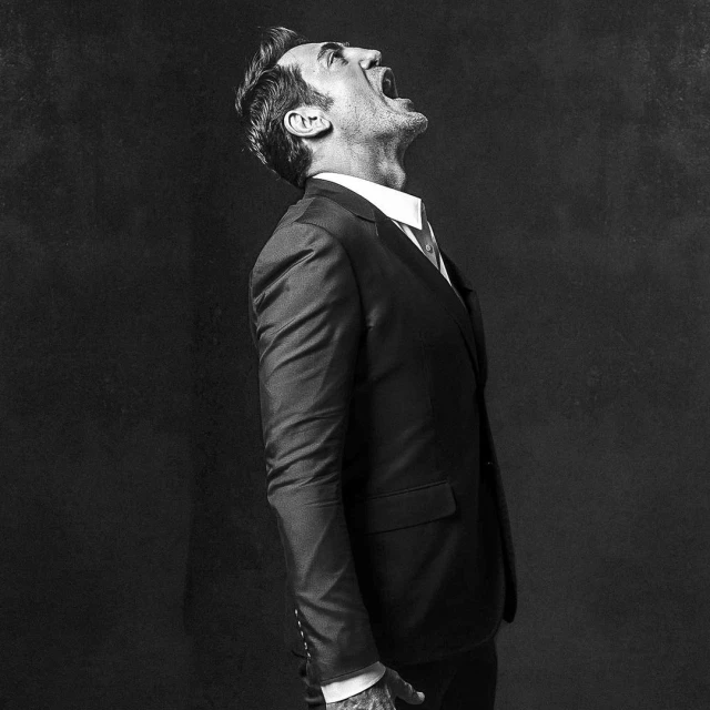 Black and white portrait of a man in a suit shouting, head tilted up, captured in a studio against a dark backdrop with natural lighting.