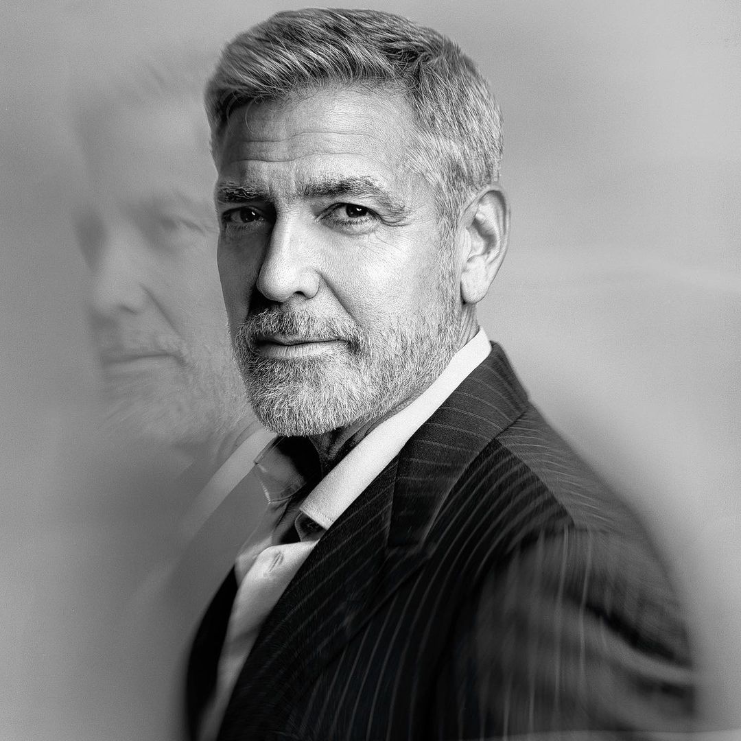 Black and white portrait of a mature adult male with short grey hair and a beard, wearing a striped suit against a soft grey background.