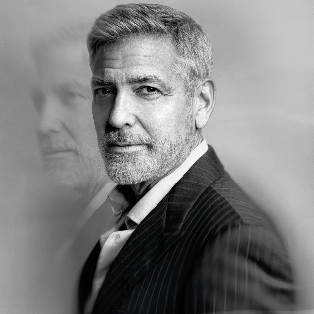 Black and white portrait of a mature adult male with short grey hair and a beard, wearing a striped suit against a soft grey background.