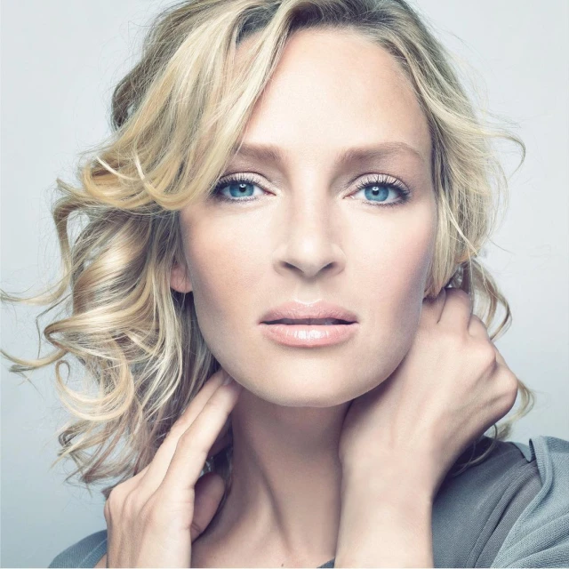 Close-up portrait of a blonde woman with blue eyes, styled wavy hair, neutral makeup, and a grey top against a pale grey background.
