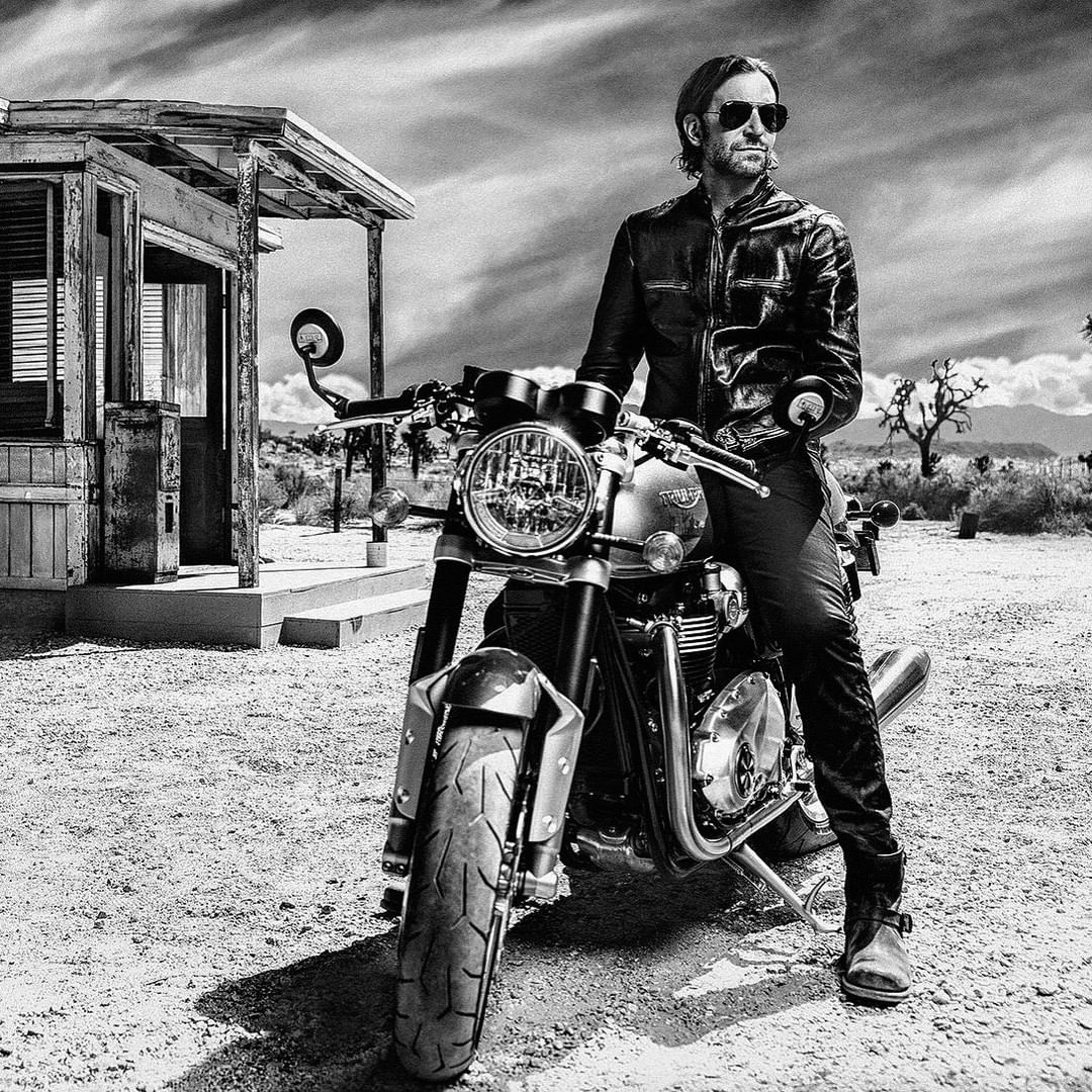 Black and white image of a man in a leather jacket sitting on a motorcycle in a desert landscape with an old building.