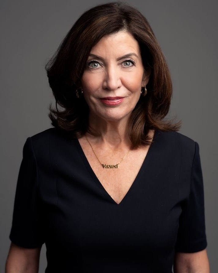 Portrait of a mature woman with shoulder-length brunette hair and wearing a black dress against a neutral grey background.