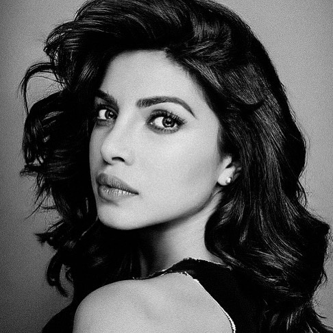 Striking black and white portrait of a South Asian adult woman with wavy dark hair, wearing dark clothing, against a neutral background.