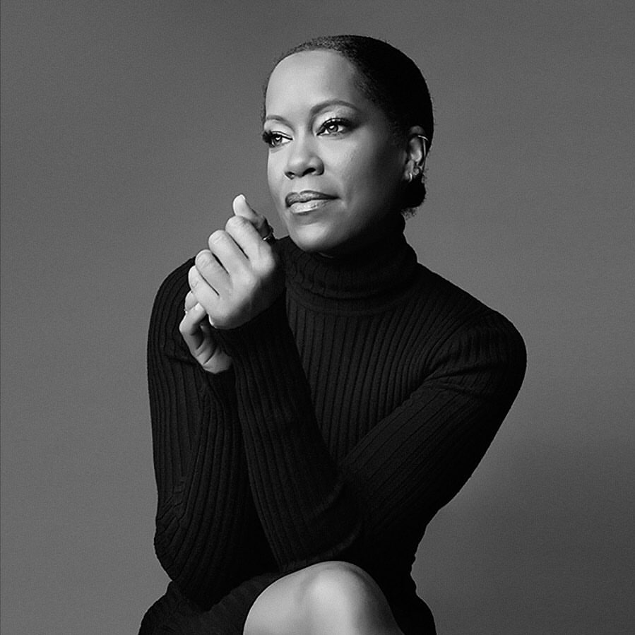 Black and white portrait of a woman with clasped hands wearing a black turtleneck sweater against a solid grey background.