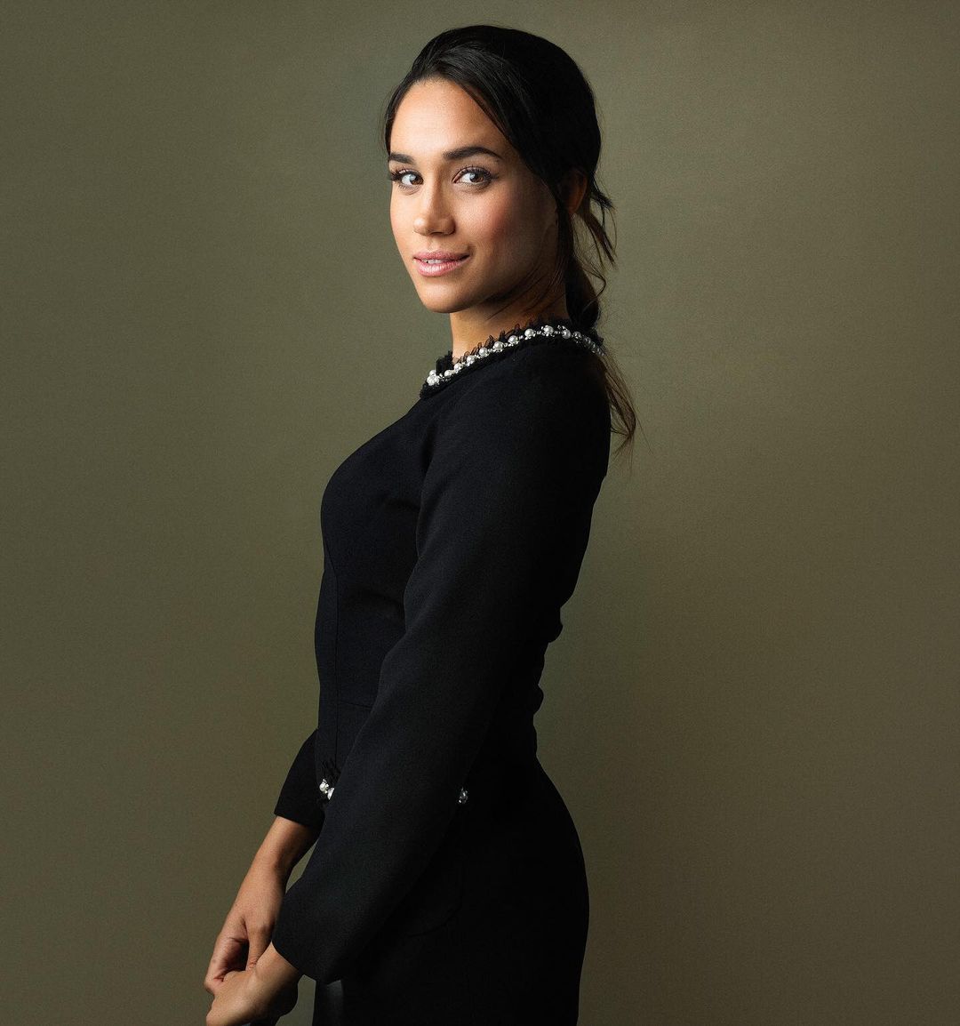 Portrait of a woman in a black long-sleeved dress with pearl detailing, looking at the camera in a studio setting with soft lighting.