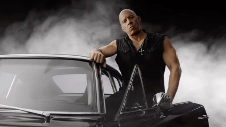 Vin Diesel leaning against a black muscle car amidst heavy white smoke, wearing a dark sleeveless shirt and a silver cross necklace.