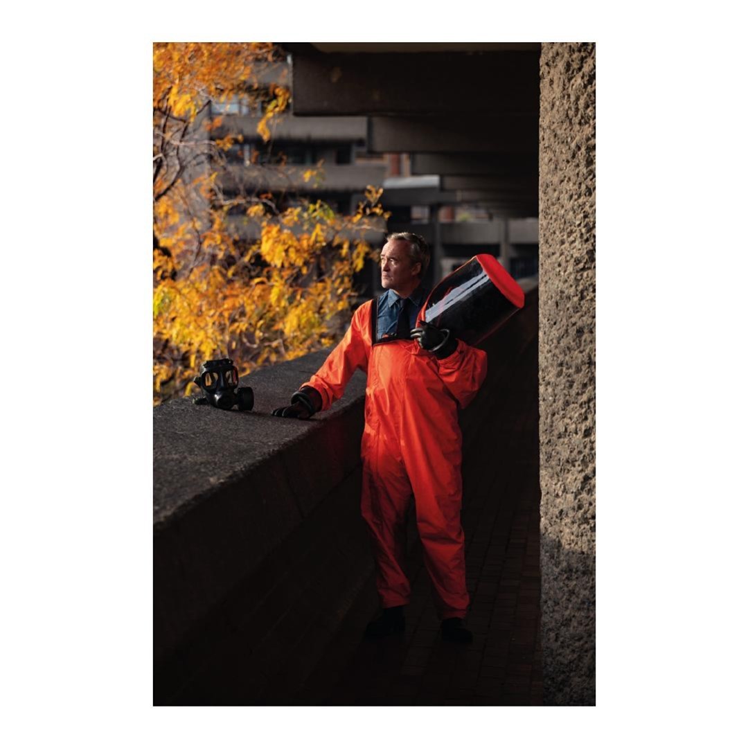 An adult male in an orange jumpsuit stands near a concrete structure, holding a dark container, with a gas mask nearby. Autumnal trees visible.