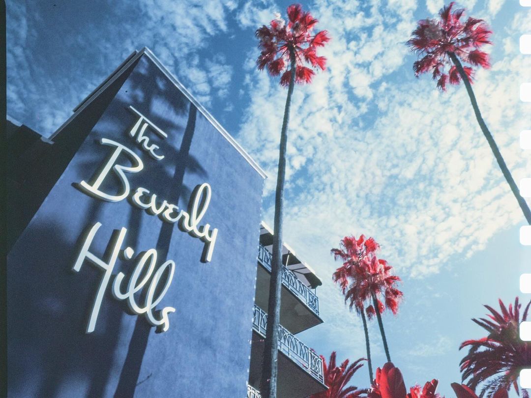 Eye-level shot of The Beverly Hills building with cursive sign in white against a blue building, palm trees, and a cloudy sky.