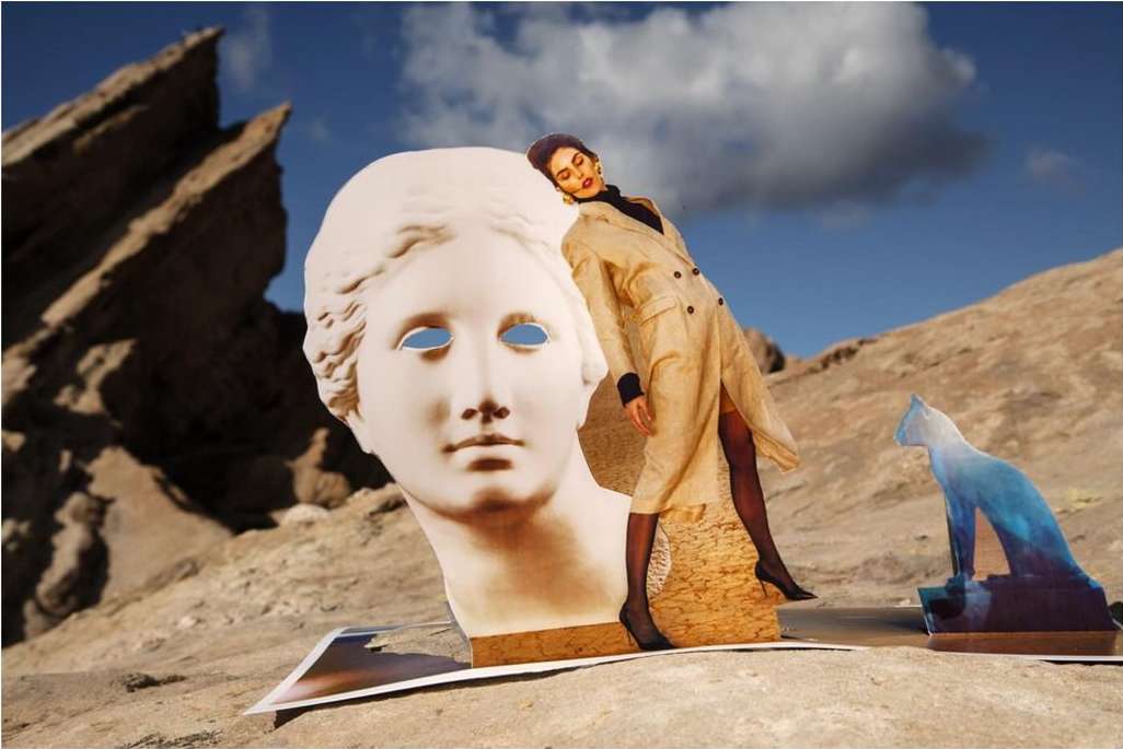 Fashion editorial in desert setting with statue head and woman in trench coat, creating surreal, artistic composition. Outdoor with blue sky.