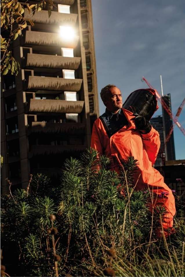An adult man carrying a large, dark bin against a high-rise apartment building under construction and lush green landscaping.