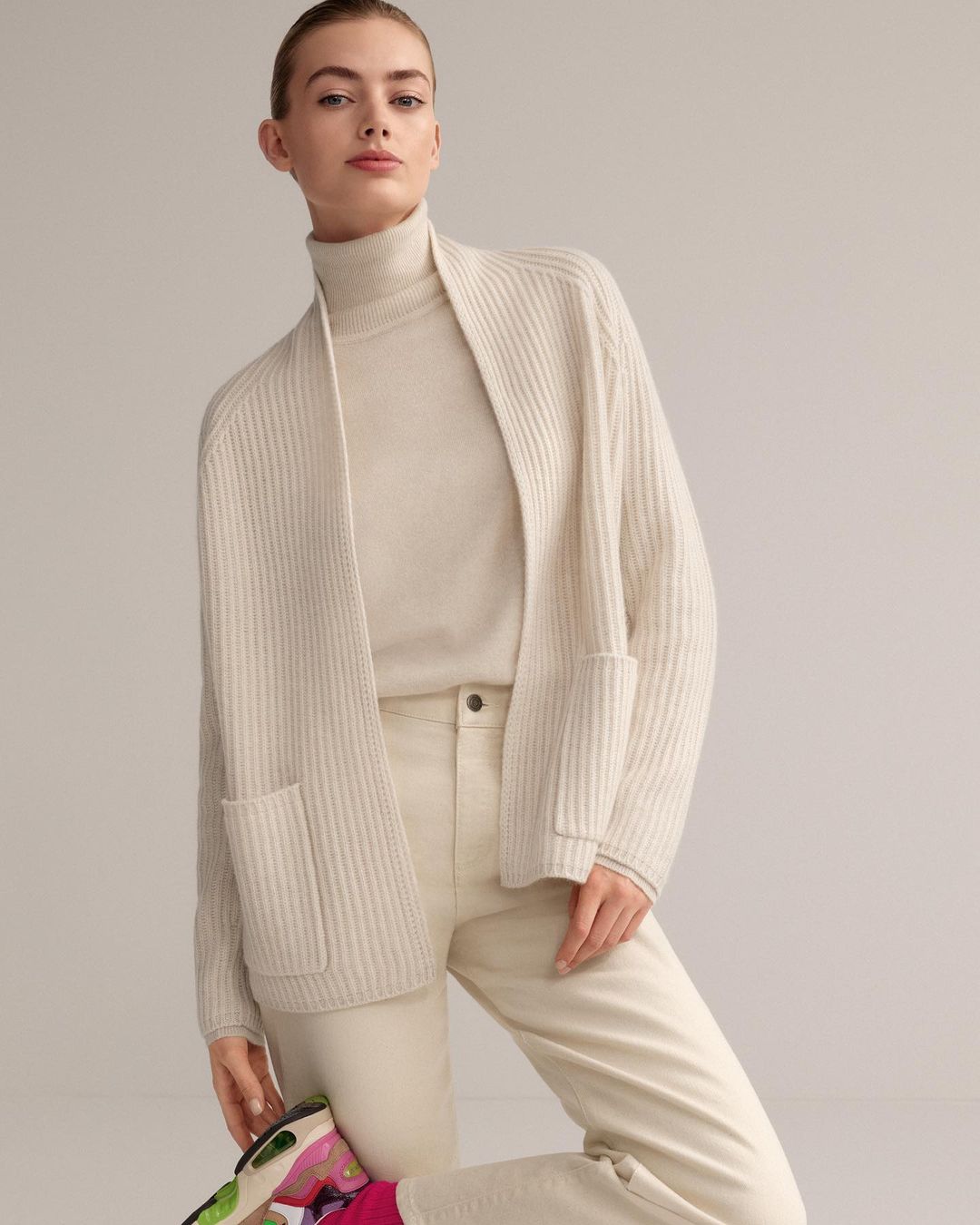 A fair-skinned woman in a casual pose, wearing a beige cardigan, turtleneck, and pants, captured in a studio setting with soft lighting.