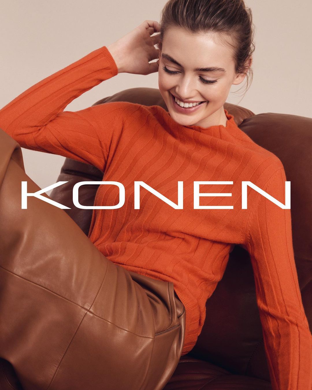 A woman in an orange ribbed sweater and brown leather pants sitting in a chair smiles while looking down in a studio shot.