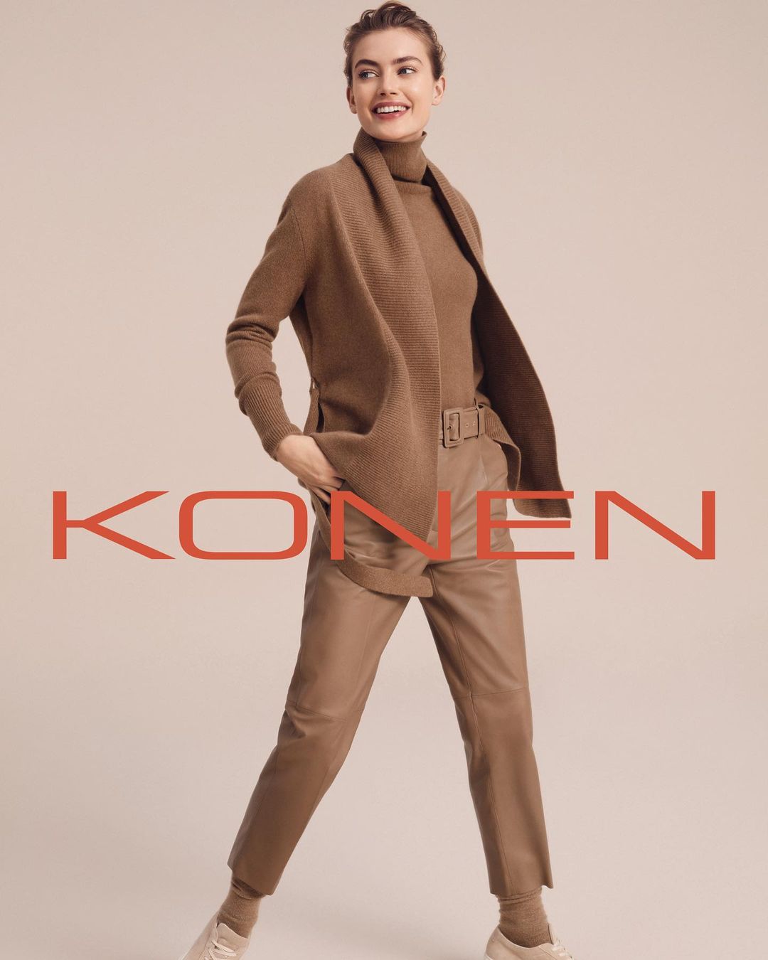 A woman in a brown turtleneck, jacket, and leather pants smiles and poses in front of a neutral background for a fashion advertisement.