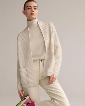 Model in cream turtleneck and ribbed cardigan with pale trousers against a solid background