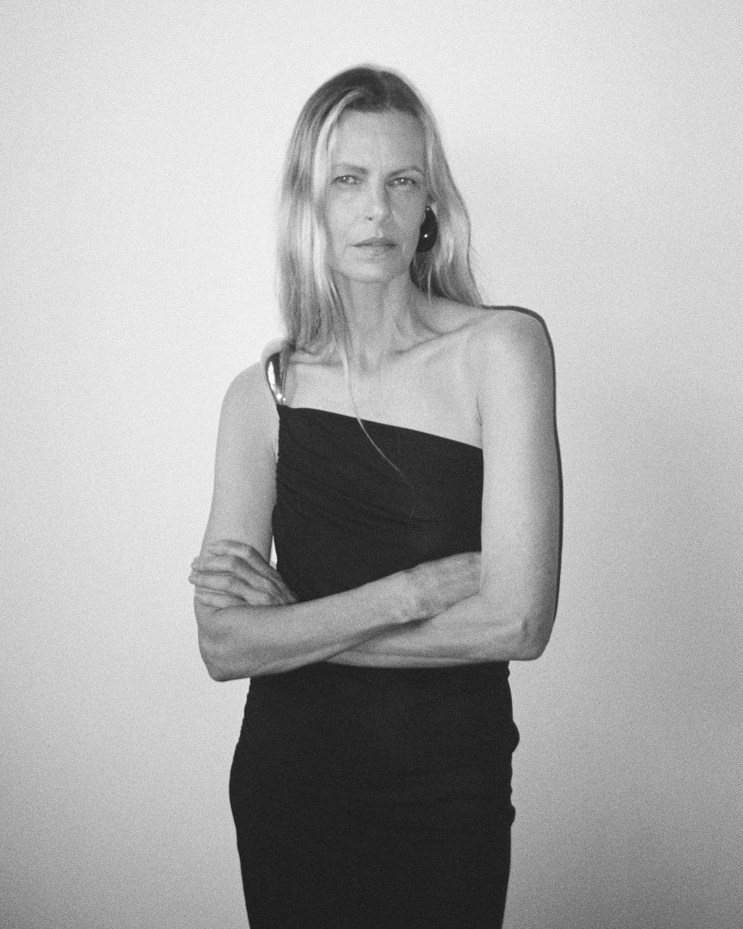 Portrait of an adult woman with long blonde hair, wearing a one-shoulder black dress, standing in front of a plain white background.