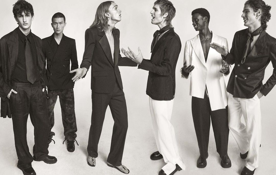 Six fashion models pose in formal wear in a black-and-white studio shot, showcasing a variety of blazers, suits, and elegant styles.
