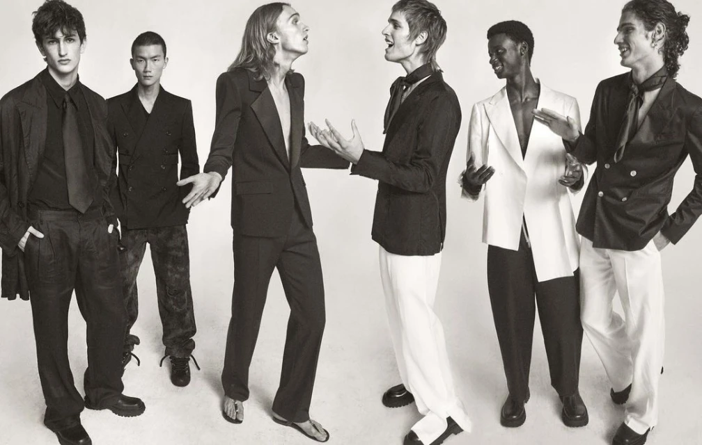 Six fashion models pose in formal wear in a black-and-white studio shot, showcasing a variety of blazers, suits, and elegant styles.