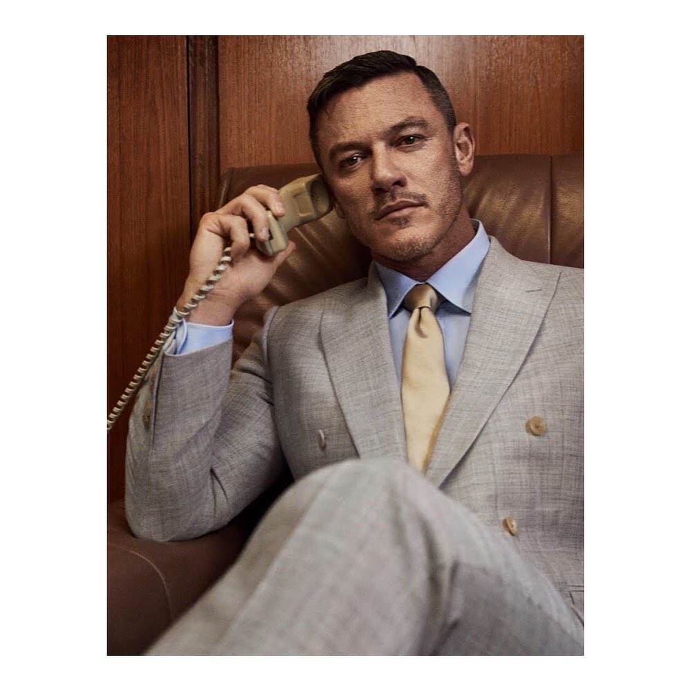 Portrait of a man in a grey suit sitting in a chair while talking on the phone in front of a wood-paneled wall. Medium shot, indoors.