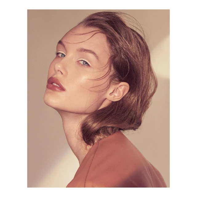 Close-up portrait of a woman with brown wavy hair, makeup with white eyeliner, wearing a dark peach top against a light tan background.
