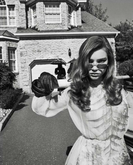 A woman in a light-colored dress and sunglasses stands in front of a stone house and driveway on a black-and-white, overcast day.