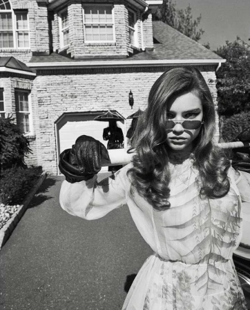 A woman in a light-colored dress and sunglasses stands in front of a stone house and driveway on a black-and-white, overcast day.