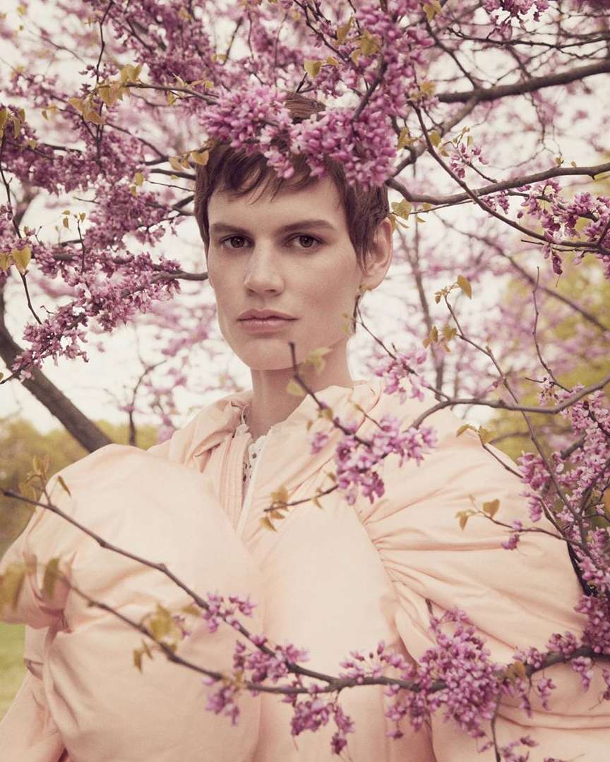 Portrait of a woman with short hair wearing a light pink dress adorned with pink flowers, framed by cherry blossoms outdoors.
