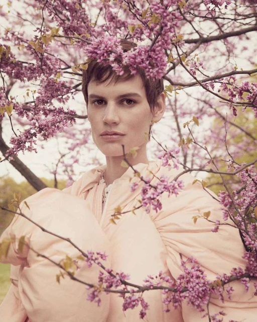 Portrait of a woman with short hair wearing a light pink dress adorned with pink flowers, framed by cherry blossoms outdoors.