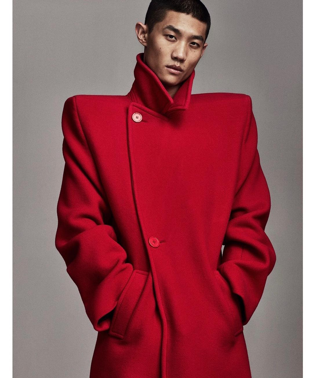 Asian male model in a red coat with strong shoulders posing in front of a plain grey background in a studio. Fashion editorial shot.