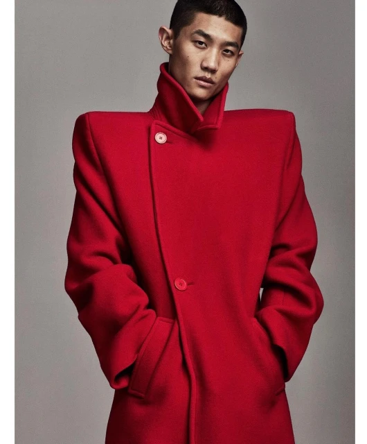 Asian male model in a red coat with strong shoulders posing in front of a plain grey background in a studio. Fashion editorial shot.