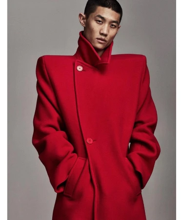 Asian male model in a red coat with strong shoulders posing in front of a plain grey background in a studio. Fashion editorial shot.