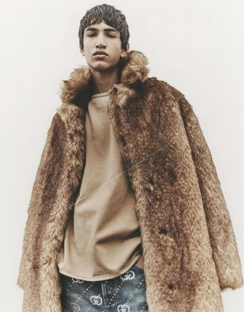 Young adult wearing a fur coat and light brown shirt stands against a plain background, showcasing a fashion-forward, upscale look.
