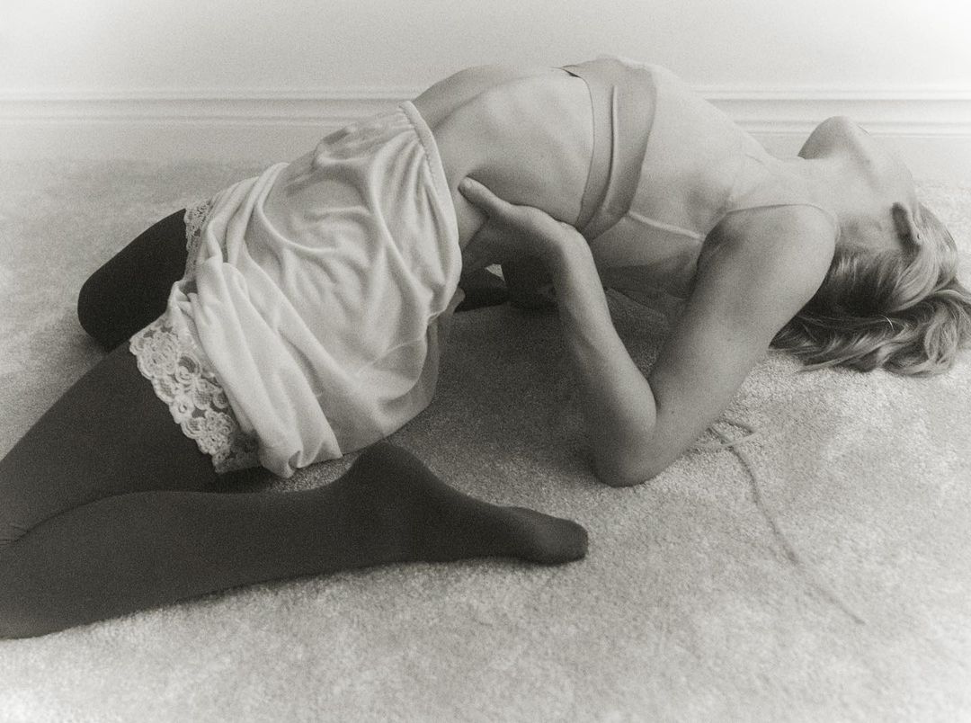 Monochrome image of a woman in a slip and tights, arched on the floor, captured in a full shot with soft, diffused light.