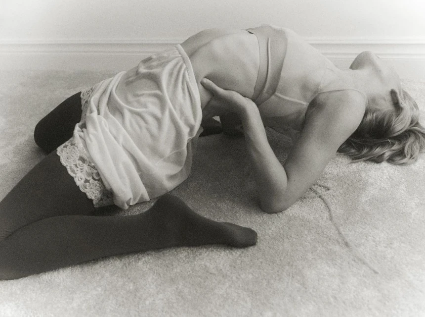 Monochrome image of a woman in a slip and tights, arched on the floor, captured in a full shot with soft, diffused light.