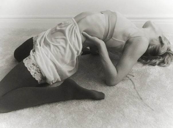 Monochrome image of a woman in a slip and tights, arched on the floor, captured in a full shot with soft, diffused light.
