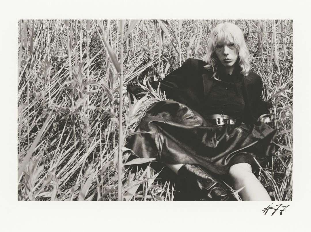 Black and white editorial portrait of a woman seated among tall grass wearing a dark jacket and belted leather skirt