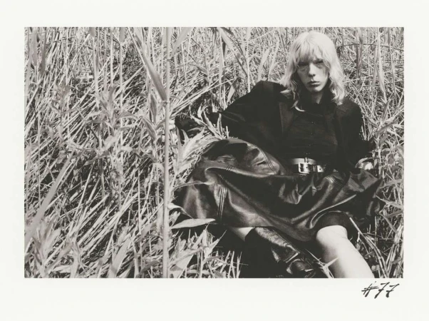 Black and white editorial portrait of a woman seated among tall grass wearing a dark jacket and belted leather skirt