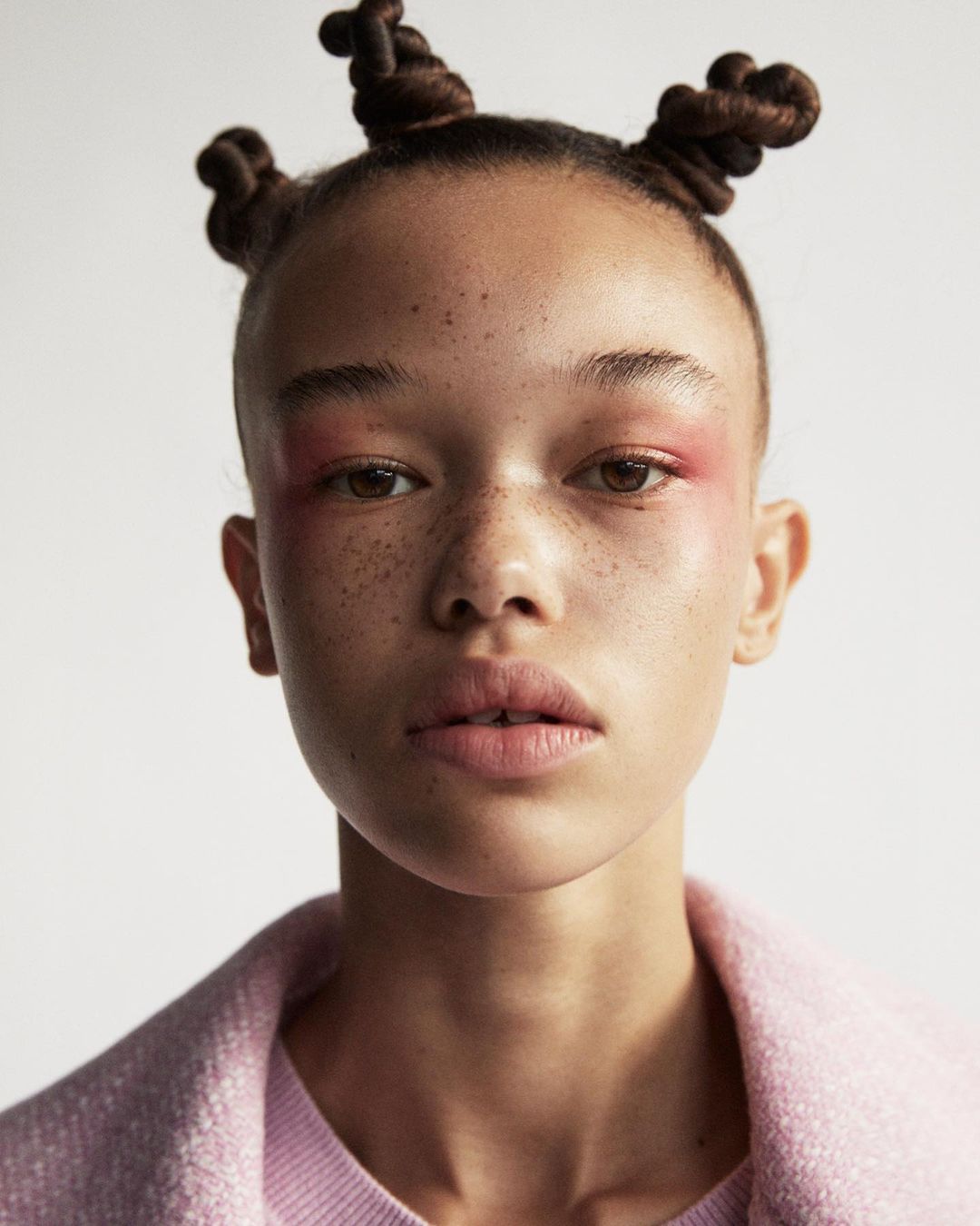 Close-up portrait of a young adult woman with freckles and creative braided hair wearing pink makeup and a pink coat against a white background.