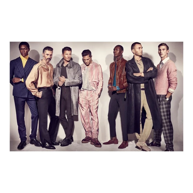 Fashion editorial shot of seven male models in various suits, jackets, pants, and coats, posing in a photo studio setting. 