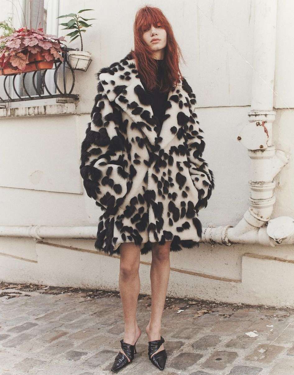A fashionable adult female stands outdoors wearing a striking black-and-white spotted faux fur coat, black top, short skirt, and stylish mules.